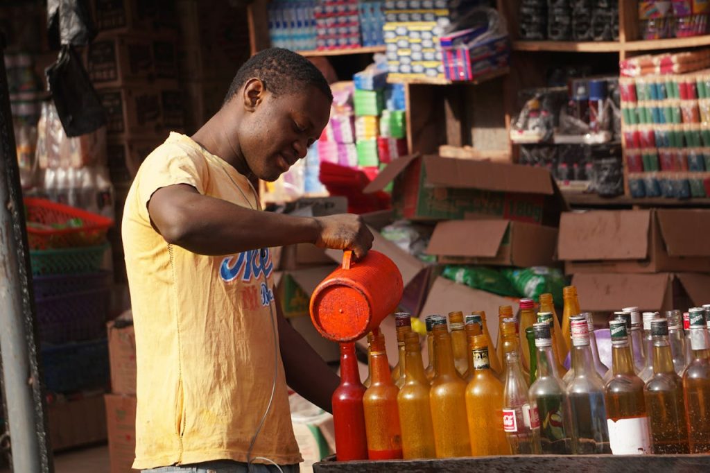 A man pouring colored liquid into several bottles at an outdoor market, surrounded by products.